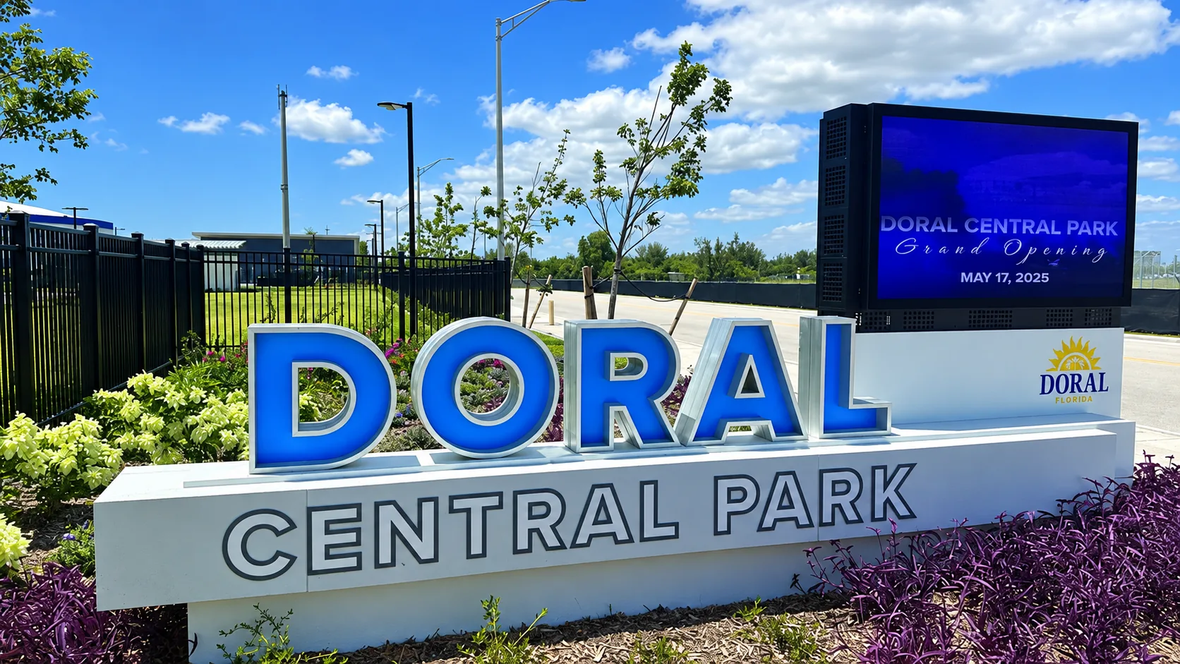 The Doral Central Park entrance sign in large blue and white lettering, surrounded by flower beds under a bright blue sky, with a digital display showing the park name beside it.