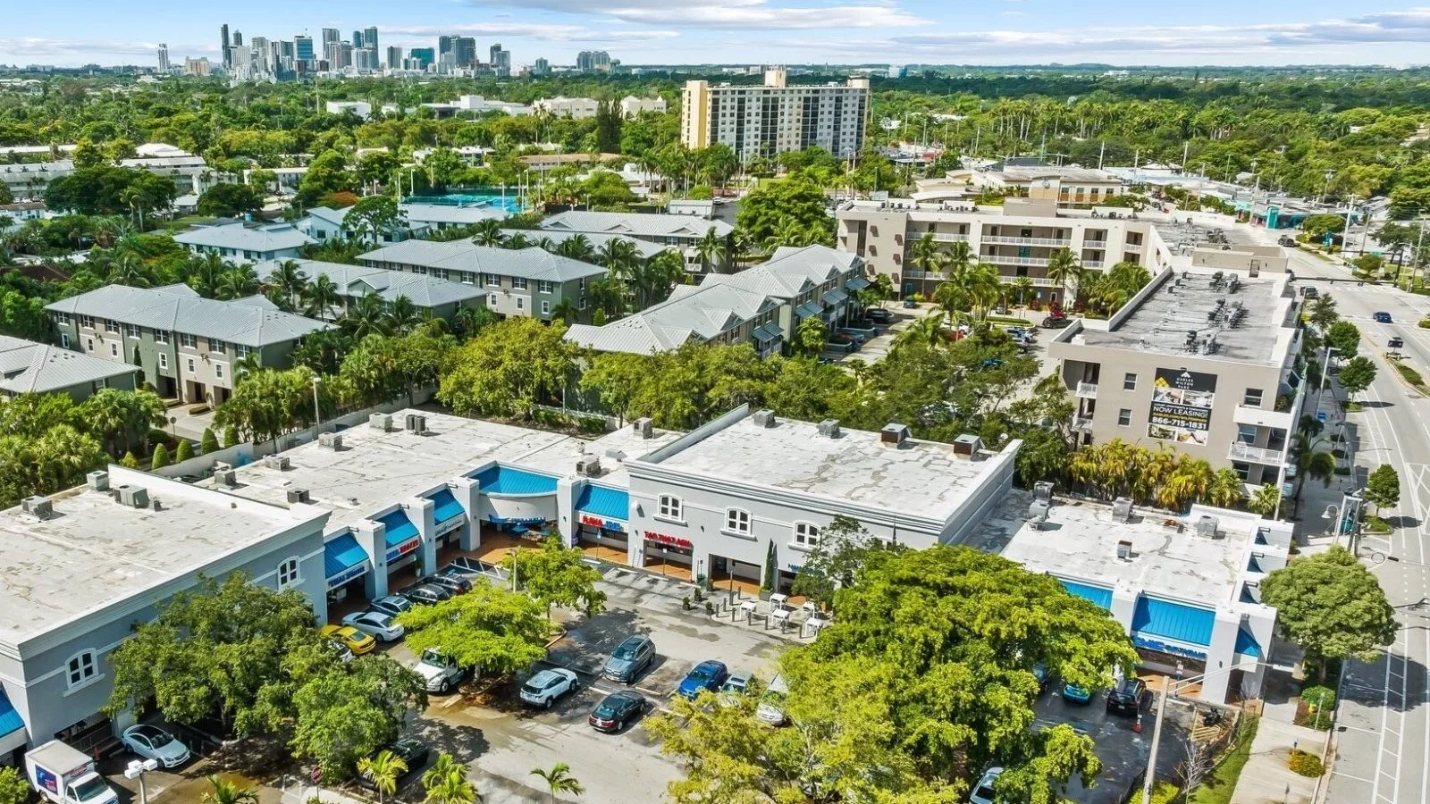 Aerial daytime view of a Florida shopping plaza with blue awnings and palm trees along Wilton Drive, with the Miami skyline visible in the distance.