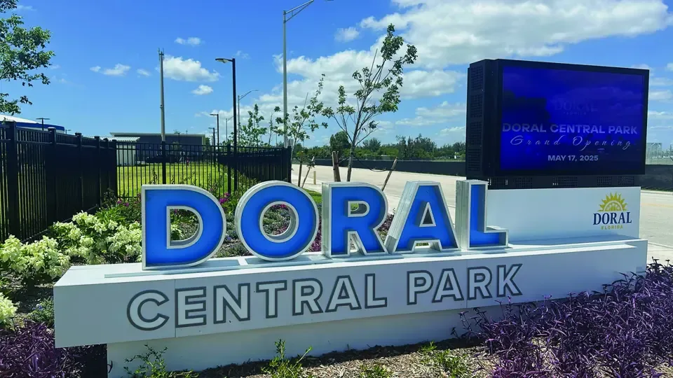 The Doral Central Park entrance sign in large blue and white lettering, surrounded by flower beds under a bright blue sky, with a digital display showing the park name beside it.