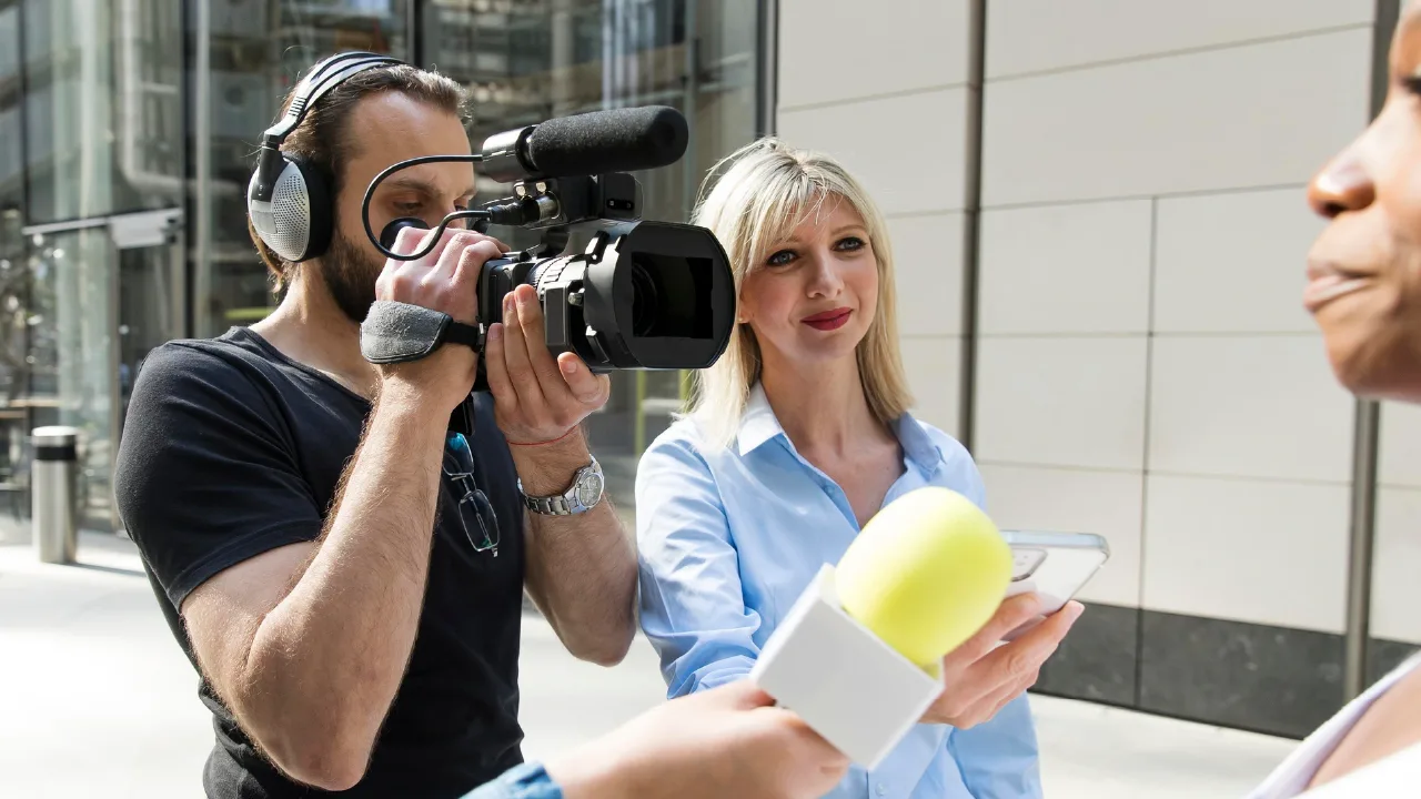 A cameraman filming with a video camera and boom microphone while a reporter interviews someone on a city street.
