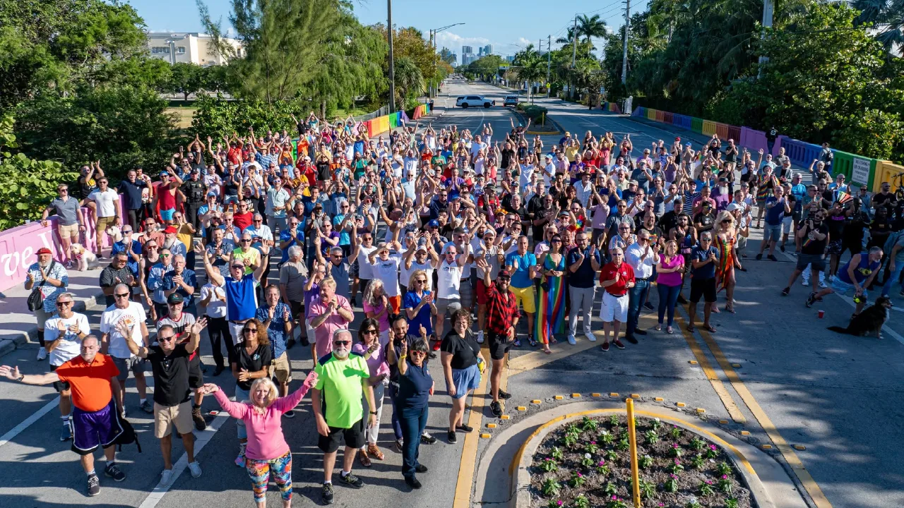 An overhead view of a large crowd filling a street in Wilton Manors, with rainbow-painted crosswalk railings and palm trees on either side.
